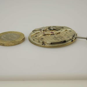 May include: A close-up of a disassembled watch movement, featuring intricate gears and a small crown, next to a gold coin. The watch mechanism is gold and silver. The coin is a gold-coloured, round coin.