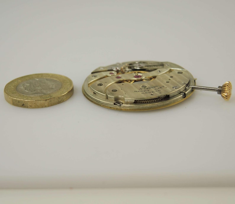 May include: A close-up of a disassembled watch movement, featuring intricate gears and a small crown, next to a gold coin. The watch mechanism is gold and silver. The coin is a gold-coloured, round coin.