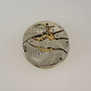 Puede incluir: Primer plano del mecanismo interno de un reloj de bolsillo de plata con engranajes dorados. La esfera del reloj est&aacute; decorada con un intrincado patr&oacute;n floral.