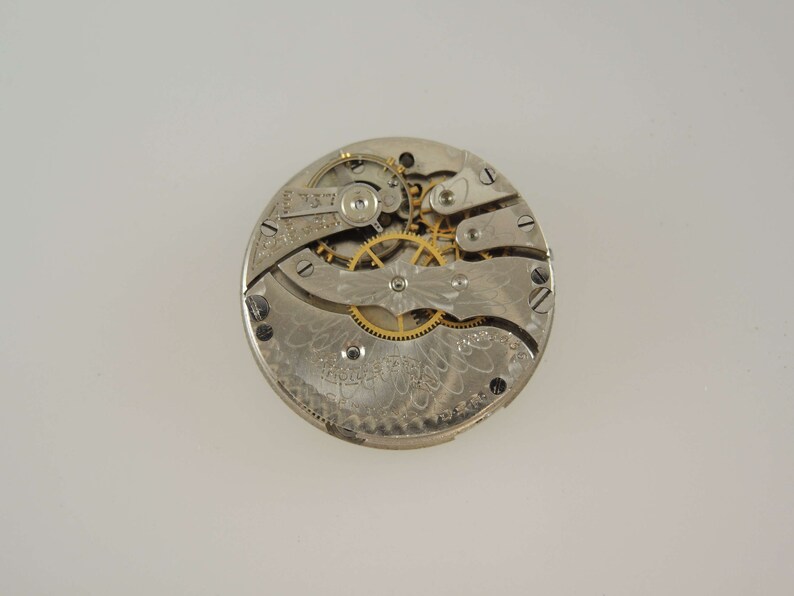 Puede incluir: Primer plano del mecanismo interno de un reloj de bolsillo de plata con engranajes dorados. La esfera del reloj est&aacute; decorada con un intrincado patr&oacute;n floral.