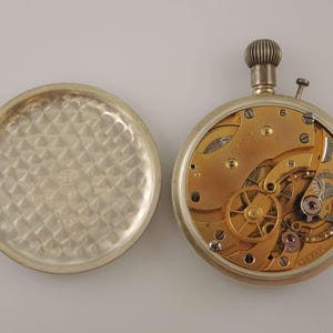 K&ouml;nnte beinhalten: Eine offene Taschenuhr, die das Innenleben zeigt. Die Uhr hat ein silbernes Geh&auml;use, ein goldfarbenes Uhrwerk mit vielen Zahnr&auml;dern und eine strukturierte Aufzugskrone. Der abgenommene R&uuml;ckdeckel hat ein strukturiertes Muster. Eine klassische Uhr.