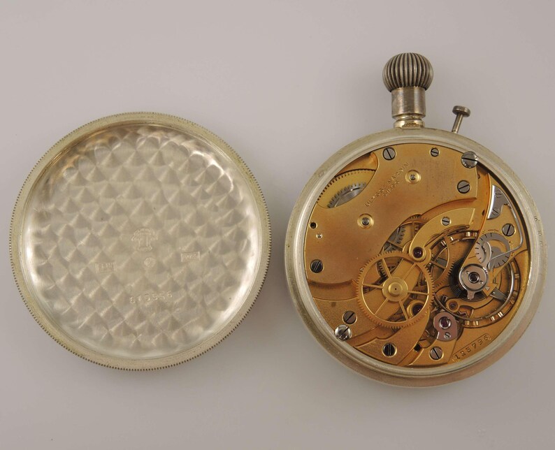 K&ouml;nnte beinhalten: Eine offene Taschenuhr, die das Innenleben zeigt. Die Uhr hat ein silbernes Geh&auml;use, ein goldfarbenes Uhrwerk mit vielen Zahnr&auml;dern und eine strukturierte Aufzugskrone. Der abgenommene R&uuml;ckdeckel hat ein strukturiertes Muster. Eine klassische Uhr.