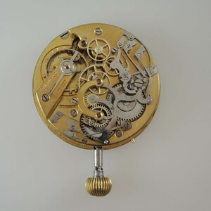 May include: A gold-toned pocket watch movement with intricate gears and a winding stem. The watch face is missing, but the intricate mechanics are visible.