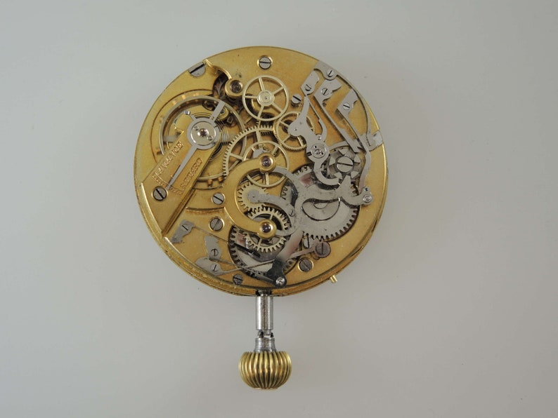 May include: A gold-toned pocket watch movement with intricate gears and a winding stem. The watch face is missing, but the intricate mechanics are visible.