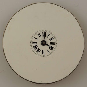May include: A white clock face with black Roman numeral markings and black hands. The clock face is surrounded by a thin gold rim and is set against a white background.