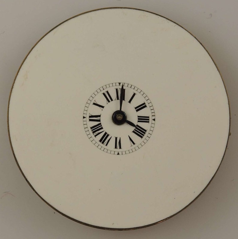 May include: A white clock face with black Roman numeral markings and black hands. The clock face is surrounded by a thin gold rim and is set against a white background.