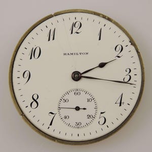 May include: A vintage Hamilton pocket watch face with a white background, black numerals, and black hands. The watch face has a subdial for seconds and the word "HAMILTON" is printed at the top.