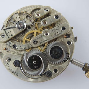 May include: Close-up of a vintage watch movement with intricate gears and mechanisms. The watch movement is silver-toned with gold-colored gears and the text "Arnold Nicoud Chaux-de-Fonds" is visible. The serial number "N&ordm; 45470" is also visible.