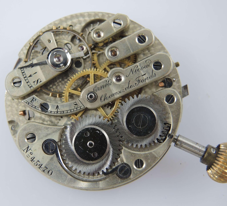May include: Close-up of a vintage watch movement with intricate gears and mechanisms. The watch movement is silver-toned with gold-colored gears and the text "Arnold Nicoud Chaux-de-Fonds" is visible. The serial number "N&ordm; 45470" is also visible.
