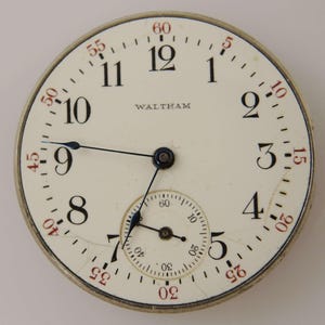 Puede incluir: Primer plano de una esfera de reloj de bolsillo Waltham vintage. La esfera blanquecina presenta números negros, marcadores de minutos rojos y un pequeño dial de segundos. Las manecillas del reloj son negras y la marca "WALTHAM" está impresa en negro.