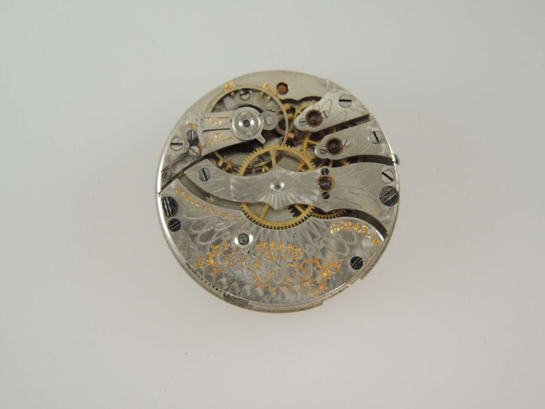 Op de afbeelding: Close-up van een zilveren zakhorloge-uurwerk met ingewikkelde tandwielen en een gouden inscriptie. De wijzerplaat van het horloge ontbreekt.