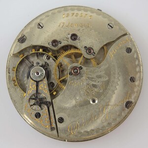 Op de afbeelding: Detailopname van een vintage zakhorlogemechanisme. Het zilverkleurige uurwerk toont goudkleurige tandwielen, schroeven en decoratieve gravures. De teksten "17 Jewels" en "Parker & Co." zijn zichtbaar. Een gedetailleerd voorbeeld van antieke horlogerie.