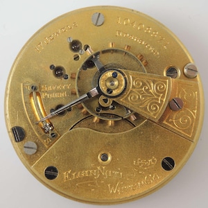 K&ouml;nnte beinhalten: Nahaufnahme eines goldfarbenen Taschenuhrwerks mit dem Text "17 Jewels" und "Elgin Natl. Watch Co." sichtbar. Das Werk hat ein dekoratives, verziertes Design.