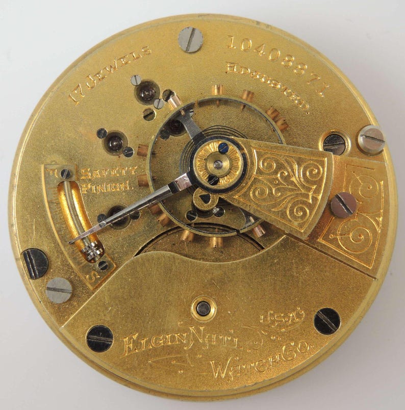 K&ouml;nnte beinhalten: Nahaufnahme eines goldfarbenen Taschenuhrwerks mit dem Text "17 Jewels" und "Elgin Natl. Watch Co." sichtbar. Das Werk hat ein dekoratives, verziertes Design.