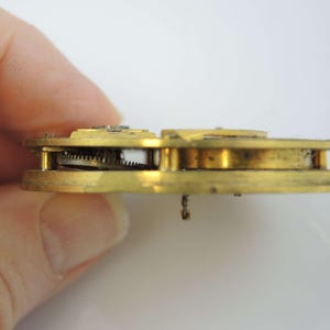 May include: Close-up side view of a gold-colored antique pocket watch movement with visible gears and a winding stem.