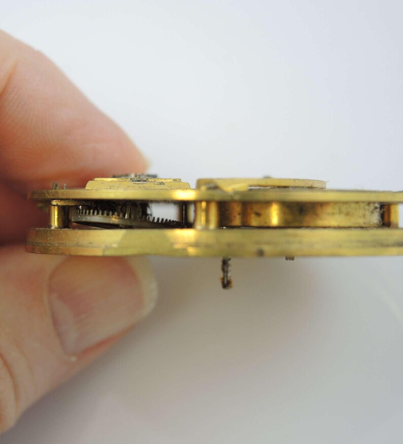 May include: Close-up side view of a gold-colored antique pocket watch movement with visible gears and a winding stem.