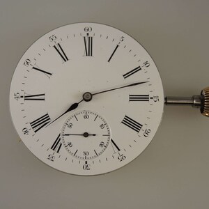 Puede incluir: Un reloj de bolsillo blanco con marcadores de hora en n&uacute;meros romanos y un subdial m&aacute;s peque&ntilde;o. El reloj tiene una llave de cuerda plateada y una perilla de color dorado.