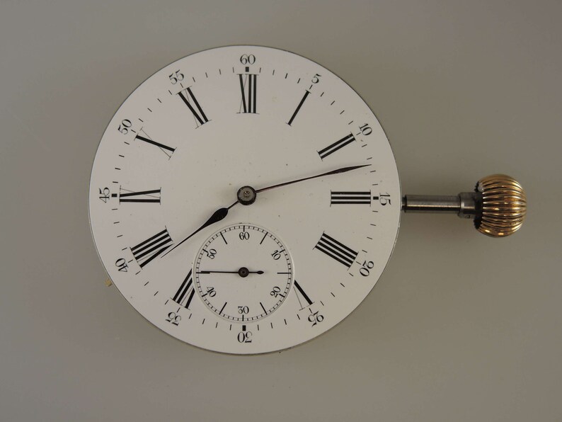 Puede incluir: Un reloj de bolsillo blanco con marcadores de hora en n&uacute;meros romanos y un subdial m&aacute;s peque&ntilde;o. El reloj tiene una llave de cuerda plateada y una perilla de color dorado.