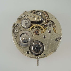 May include: Close-up of a silver pocket watch movement with intricate gears and a winding stem. The watch face is not visible.