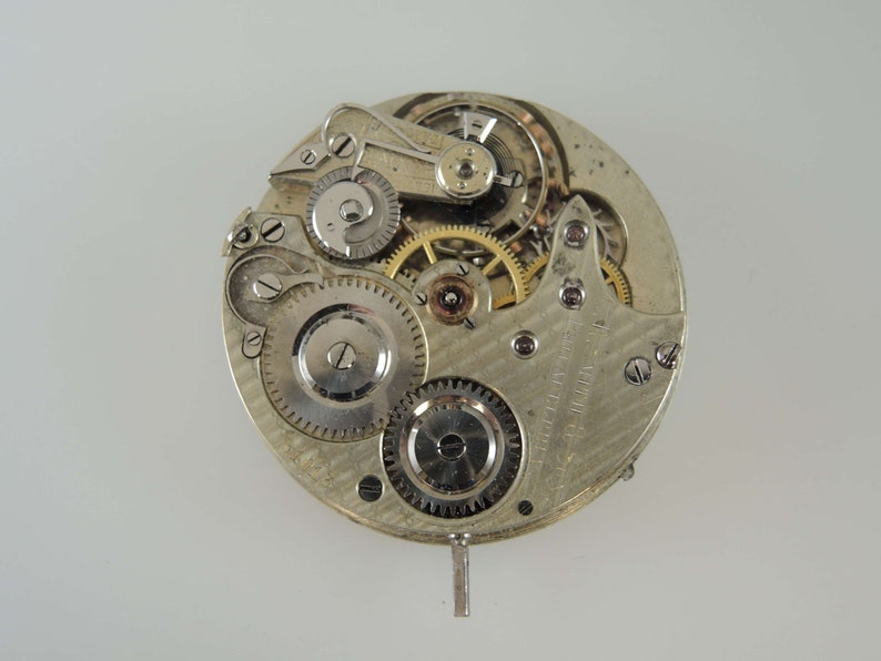 May include: Close-up of a silver pocket watch movement with intricate gears and a winding stem. The watch face is not visible.