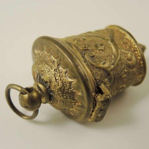 May include: A small, gold-colored, ornate, hinged, cylindrical box with a loop for hanging. The box has a detailed, embossed design.
