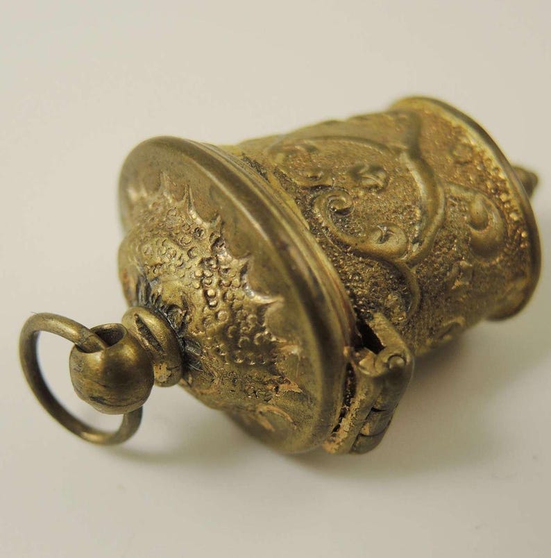 May include: A small, gold-colored, ornate, hinged, cylindrical box with a loop for hanging. The box has a detailed, embossed design.