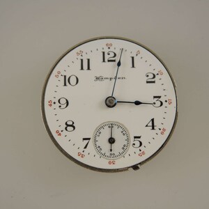 Op de afbeelding: Een wit zakhorloge met zwarte wijzers en zwarte Romeinse cijfers voor de uren. Het horloge heeft een kleinere, concentrische cirkel met zwarte markeringen voor de seconden. Het horloge heeft ook de tekst "Hamilton" erop gedrukt.