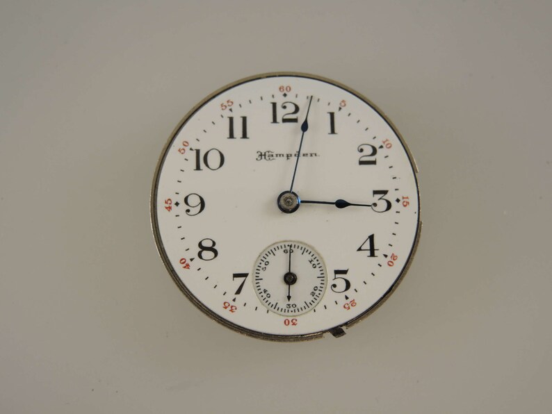 Op de afbeelding: Een wit zakhorloge met zwarte wijzers en zwarte Romeinse cijfers voor de uren. Het horloge heeft een kleinere, concentrische cirkel met zwarte markeringen voor de seconden. Het horloge heeft ook de tekst "Hamilton" erop gedrukt.