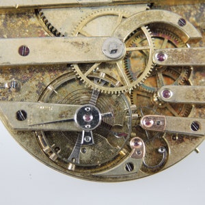 May include: Detailed view of a vintage watch mechanism, revealing numerous small gears, levers, and screws. The metal parts have a weathered, aged look, with tiny ruby jewels set in place. The design is circular, featuring a complex arrangement of mechanical components.