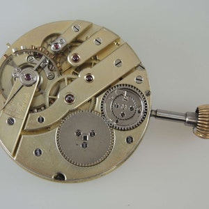 Puede incluir: Una vista cercana del funcionamiento interno de un movimiento de reloj de bolsillo dorado. El reloj tiene un v&aacute;stago de cuerda y una variedad de engranajes y resortes.