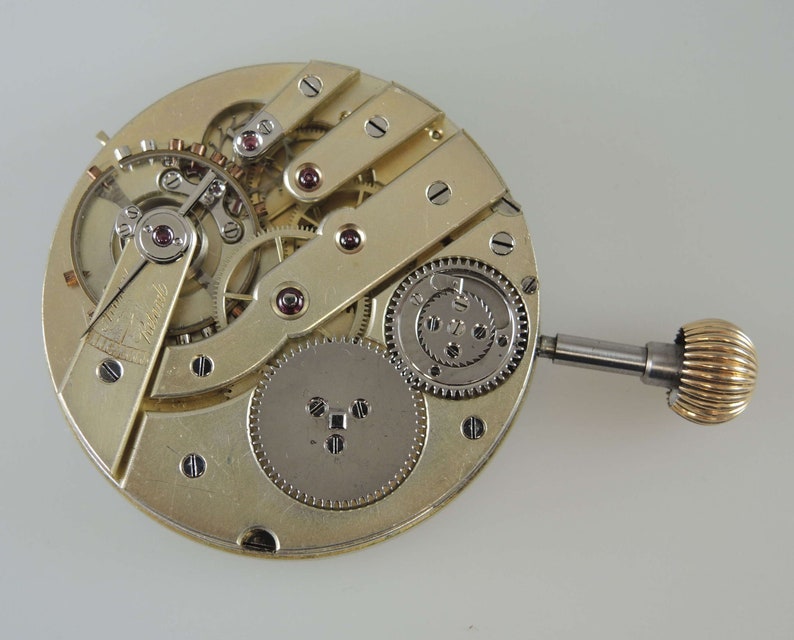 Puede incluir: Una vista cercana del funcionamiento interno de un movimiento de reloj de bolsillo dorado. El reloj tiene un v&aacute;stago de cuerda y una variedad de engranajes y resortes.