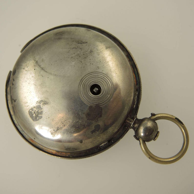 May include: A vintage silver pocket watch with a circular design. The watch has a tarnished appearance, with a small, decorative circle near the center. A gold-colored ring is attached to the side.