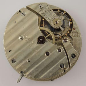 May include: Close-up of a vintage watch movement with intricate gears and components. The silver-toned metal base features engraved details and the words "FAST" and "SLOW". The watch mechanism is a detailed example of horological engineering.