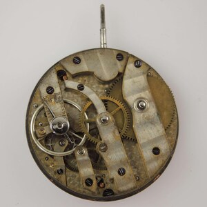 May include: Close-up of a vintage watch movement, showcasing intricate gears, levers, and screws. The metal components have a worn, aged appearance, with a circular design and a winding stem at the top. The watch mechanism is a testament to precision engineering.