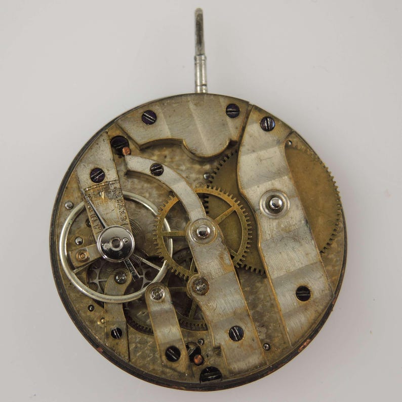 May include: Close-up of a vintage watch movement, showcasing intricate gears, levers, and screws. The metal components have a worn, aged appearance, with a circular design and a winding stem at the top. The watch mechanism is a testament to precision engineering.