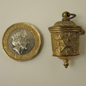 May include: A small, gold-colored, hinged, decorative container with an ornate design. The container is about the size of a British one pound coin.