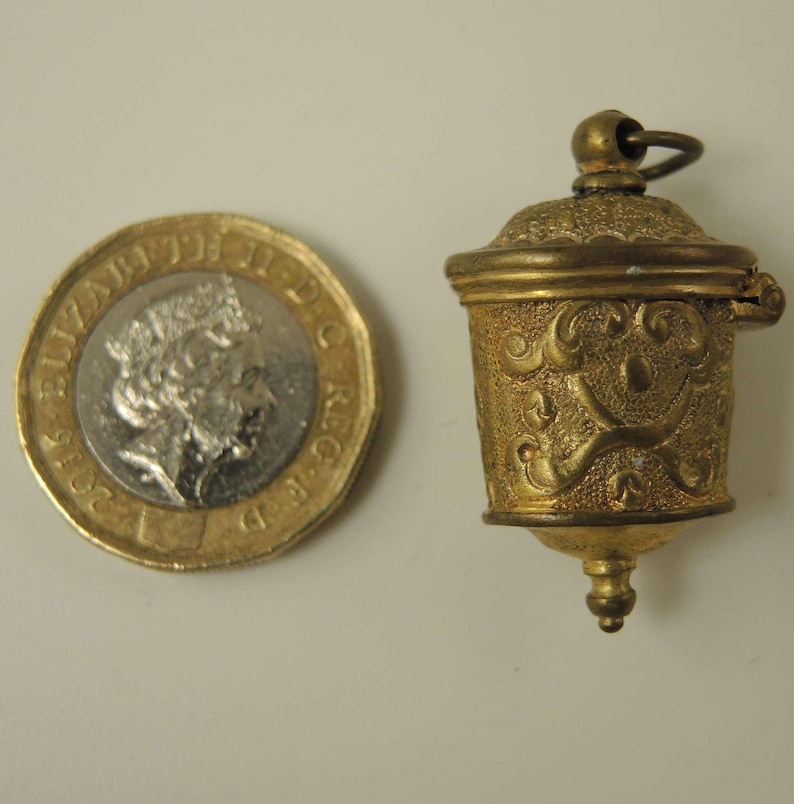 May include: A small, gold-colored, hinged, decorative container with an ornate design. The container is about the size of a British one pound coin.