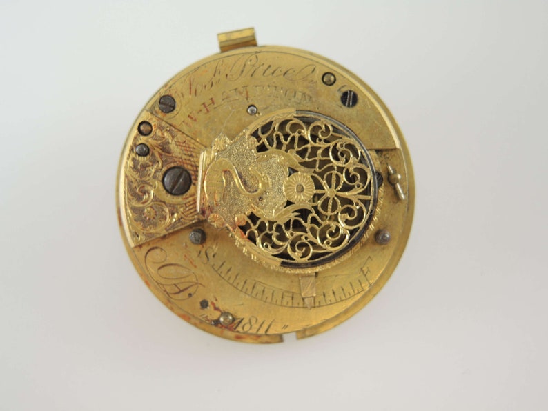 K&ouml;nnte beinhalten: Nahaufnahme eines Vintage-Taschenuhrwerks. Der goldfarbene Mechanismus weist komplizierte Details auf, darunter Zahnr&auml;der, Schrauben und eine dekorative Filigranarbeit. Die Uhr ist mit Text und dem Jahr 1811 beschriftet.