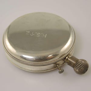K&ouml;nnte beinhalten: Eine silberfarbene, runde Taschenuhr mit strukturiertem Aufzugsknopf und kleinem Knopf. Die Uhr hat die eingravierten Initialen "H.S.12" auf dem Deckel. Die Uhr ist ein Vintage-Artikel.