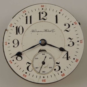 May include: A white pocket watch face with black Roman numeral hour markers and black hands. The watch face has a smaller, concentric circle with black markings for seconds. The watch face also has the text "Hampden Watch Co." printed on it.