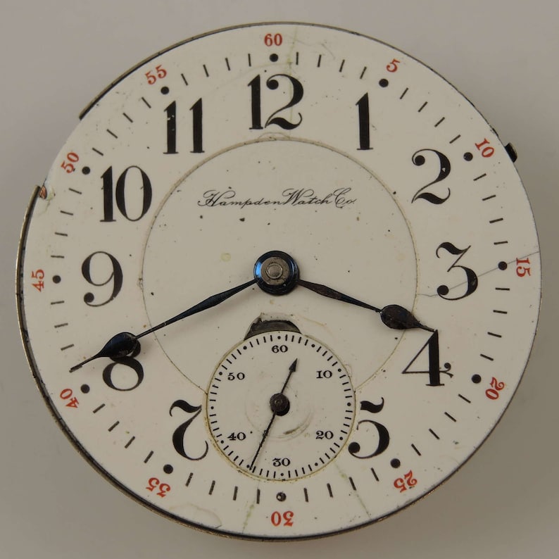 May include: A white pocket watch face with black Roman numeral hour markers and black hands. The watch face has a smaller, concentric circle with black markings for seconds. The watch face also has the text "Hampden Watch Co." printed on it.