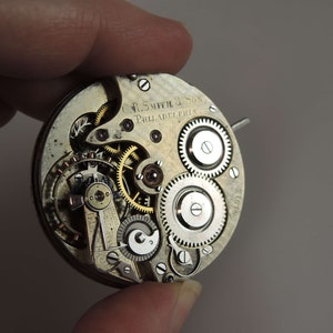 May include: Close-up of a silver pocket watch movement with intricate gears and the inscription "C.F. Smith & Son, Philadelphia".