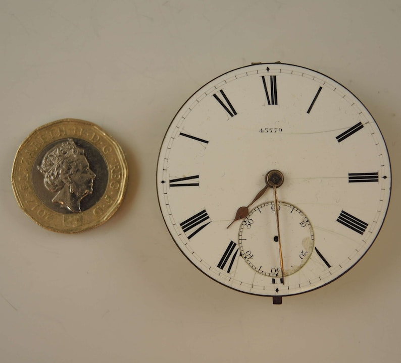 May include: A white pocket watch face with black Roman numeral hour markers and black minute markers. The watch face has a small seconds dial at the 6 o'clock position. The watch face has the number 45779 printed on it.