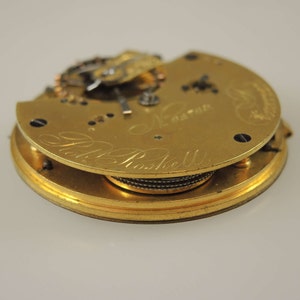 May include: Close-up of a gold pocket watch movement with the inscription "Robt. Roskell" and "London" visible. The watch is disassembled and the inner workings are exposed.