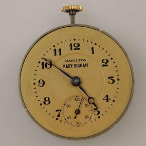 K&ouml;nnte beinhalten: Antikes Taschenuhrzifferblatt mit cremefarbenem Zifferblatt, schwarzen Ziffern und blauen Zeigern. Das Zifferblatt tr&auml;gt den Text "HAMILTON MARY BIGHAM". Die Uhr hat eine kleine Sekundenscheibe und eine goldfarbene Aufzugswelle.