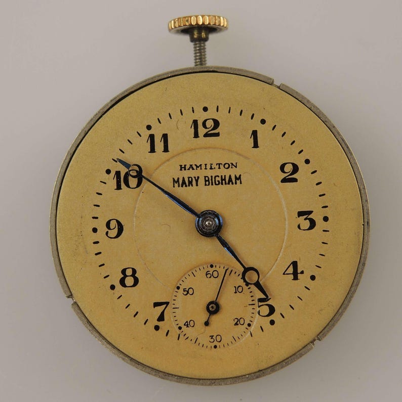 K&ouml;nnte beinhalten: Antikes Taschenuhrzifferblatt mit cremefarbenem Zifferblatt, schwarzen Ziffern und blauen Zeigern. Das Zifferblatt tr&auml;gt den Text "HAMILTON MARY BIGHAM". Die Uhr hat eine kleine Sekundenscheibe und eine goldfarbene Aufzugswelle.