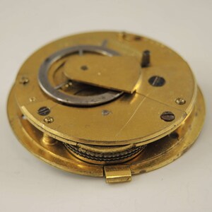 May include: A gold-toned pocket watch mechanism with a circular face and a hinged cover. The watch face has a silver-toned gear and a smaller circular gear. The watch mechanism is open, revealing the inner workings.
