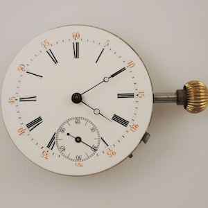 May include: A vintage watch face with a white background, black Roman numerals, and a smaller seconds dial. The minute markers are in orange. The watch has a silver stem and a gold-colored crown.