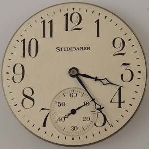 Könnte beinhalten: Eine weiße Taschenuhr mit schwarzen Zeigern und schwarzen römischen Ziffern für die Stunden. Das Zifferblatt der Uhr hat ein kleineres, kreisförmiges Hilfszifferblatt mit schwarzen arabischen Ziffern. Das Zifferblatt der Uhr trägt auch den Schriftzug "STUDEBAKER".