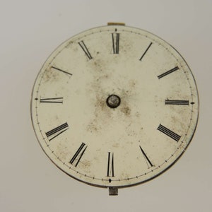 May include: A white clock face with black Roman numeral hour markers and a black minute track. The clock face is slightly dirty and has a small metal knob in the center.